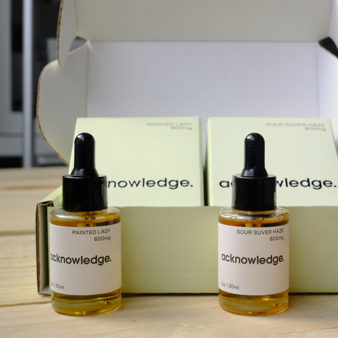 A photo featuring the full suite of Acknowledge packaging—a mailer box, two different product boxes, and two bottles. These products are sitting on a wooden board.