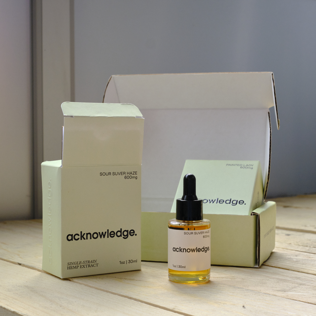 A photo featuring the full suite of Acknowledge packaging—a mailer box, two different product boxes, and a bottle. These products are sitting on a wooden board.