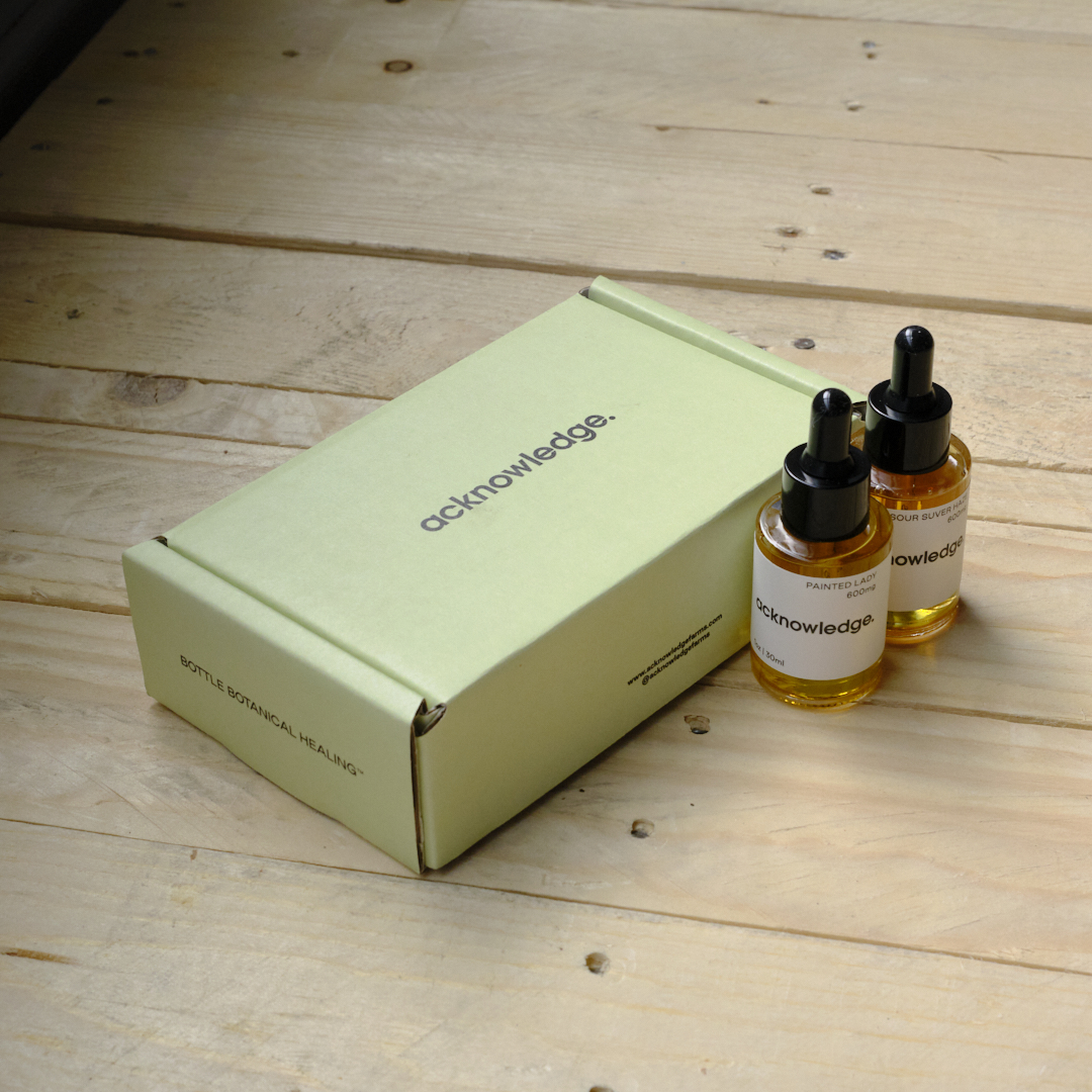A photo featuring the full suite of Acknowledge packaging—a mailer box and two bottles. These products are sitting on a wooden board.