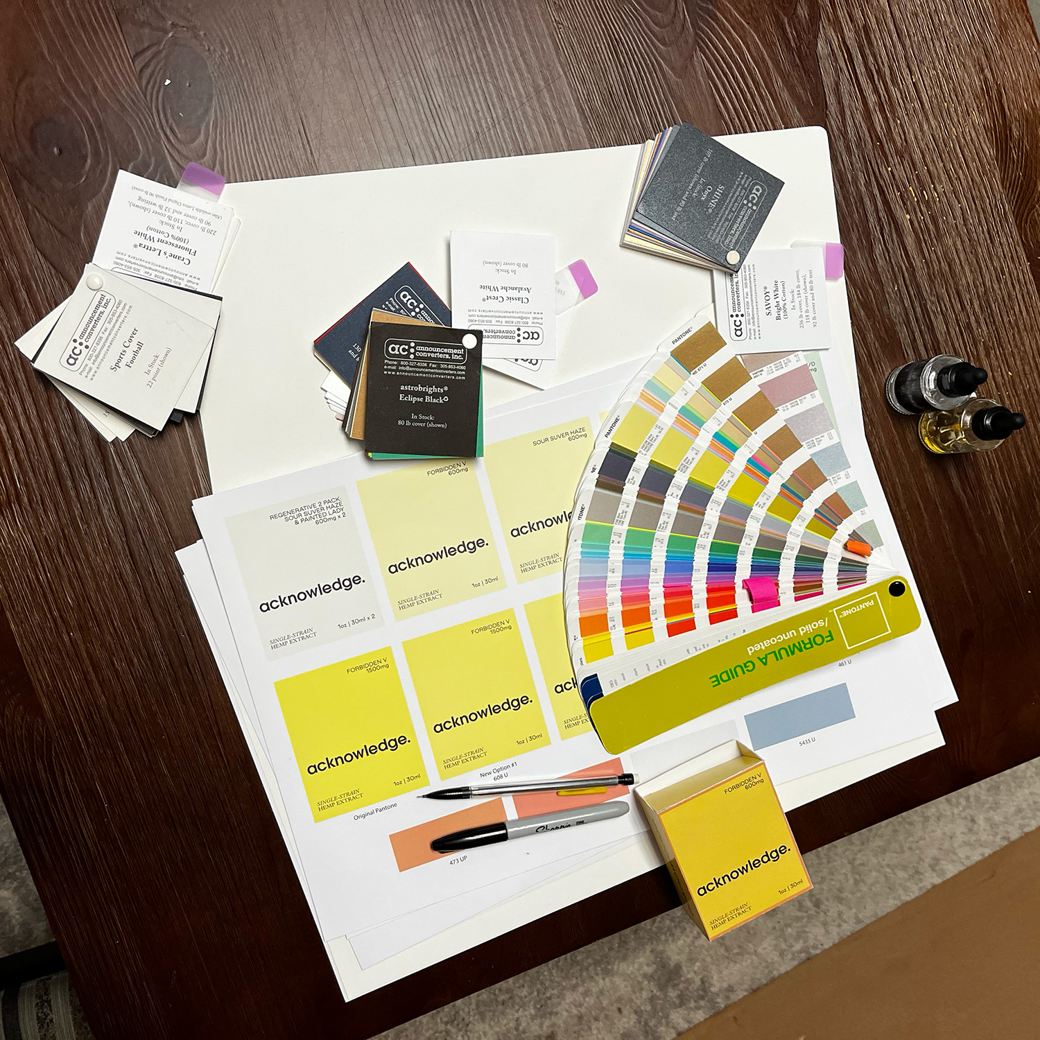 A somewhat messy table featuring many different types of paper, a Pantone color matching book, and a few rough prototypes of packaging.