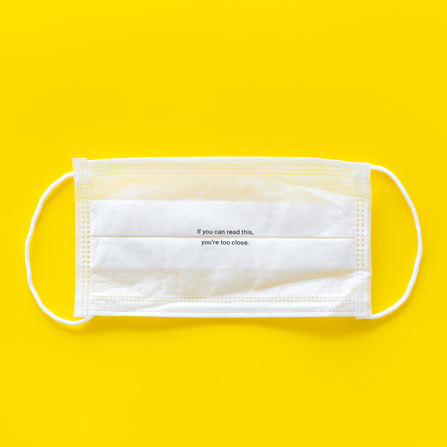 A white facemask, placed on a stark yellow backdrop. There is very small text on the front of the mask, which is illegible from a distance. When you get close enough to read it, you can read it, and it says 'If you can read this, you're too close.'