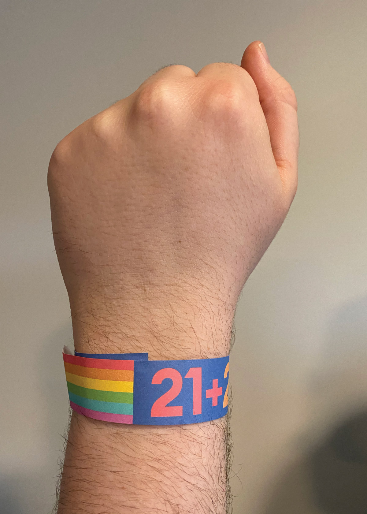 A photo showing what a twenty-one plus barhopping wristband might look like. I printed and wrapped this around a real wrist for this images, and the wrist is pointing upward in a 'resistance' pose. The wristband is navy blue, with the numbers twenty-one and a plus sign repeating across the band in various rainbow colors. The section over the taped part of the band is decorated as a pride flag.